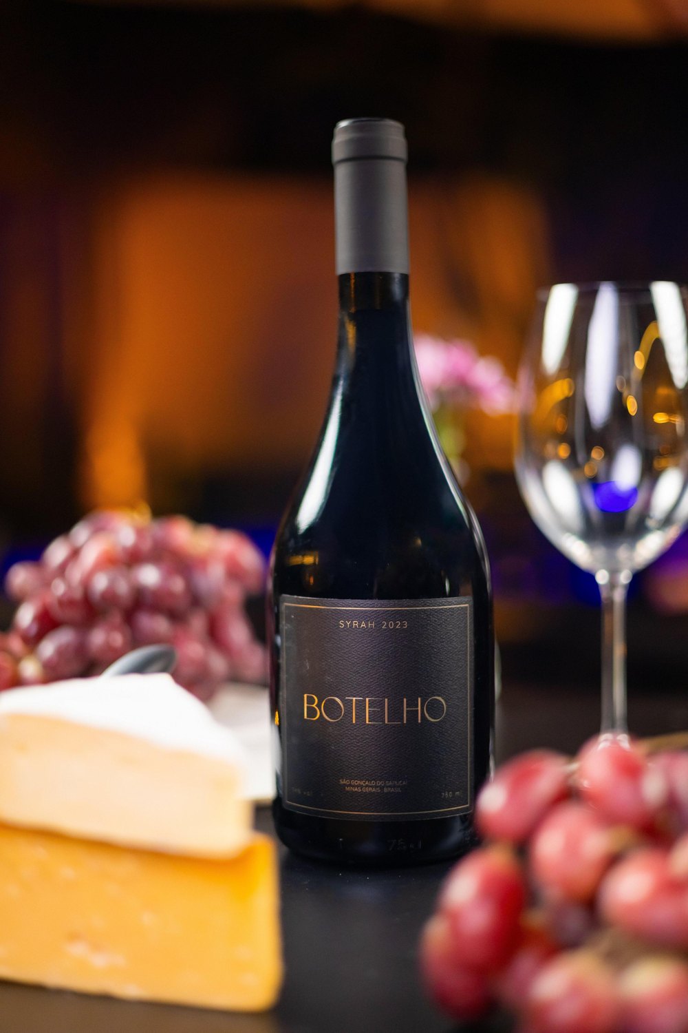 Botelho Syrah 2023 sobre a mesa, com queijos, uvas e uma taça